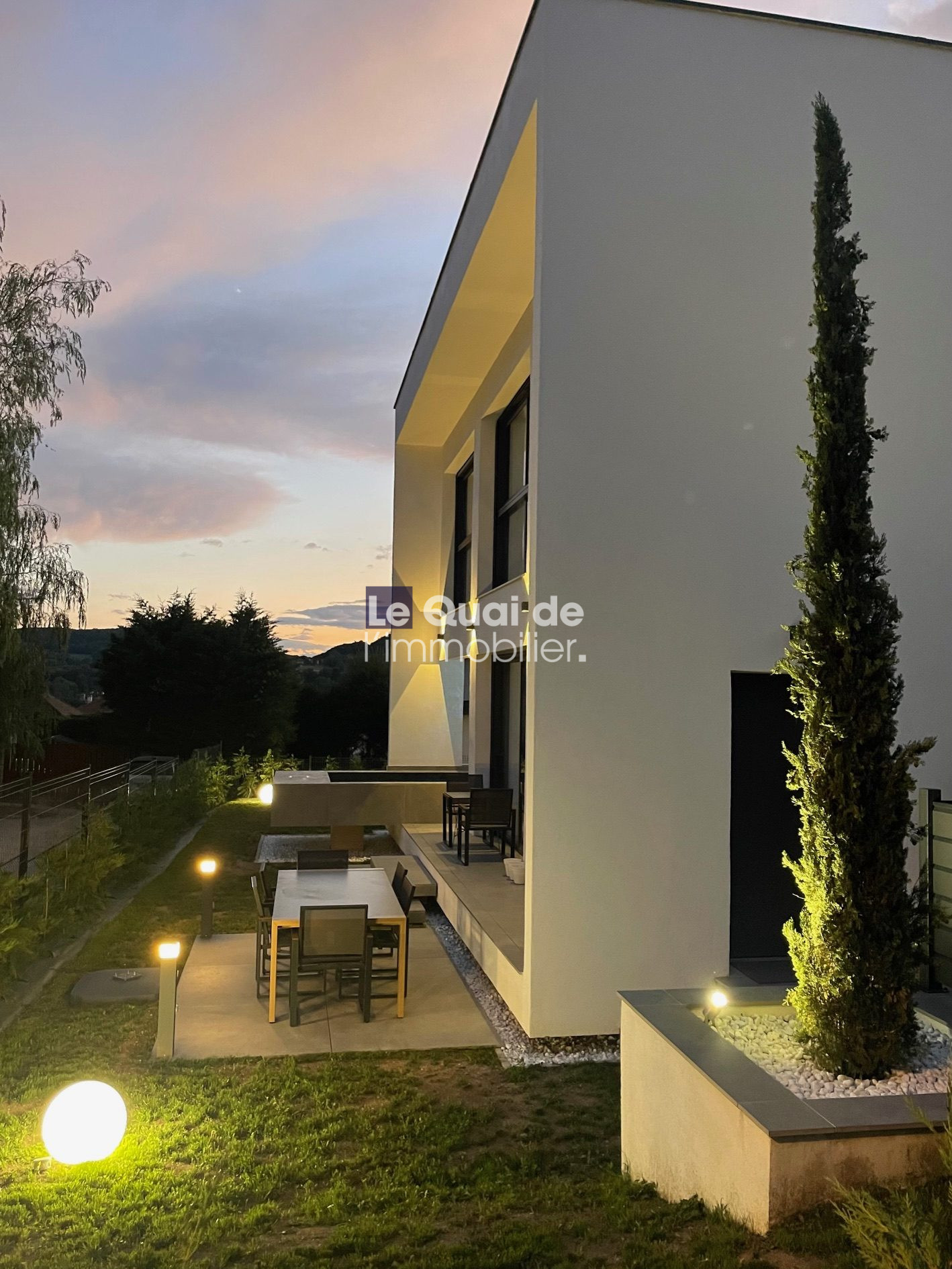 Modern white two‑story house with a lit patio, outdoor dining table and chairs on a tiled terrace, a grassy lawn with pathway lights, and a tall slender tree by the entrance.