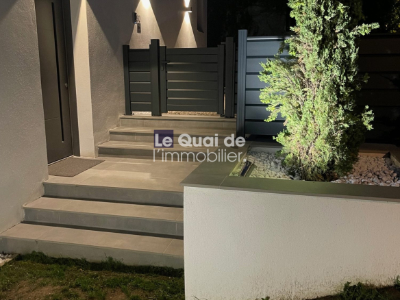 Night exterior entrance: illuminated wall sconces cast triangular light patterns on a gray wall beside a dark door and steps leading to a black gate.