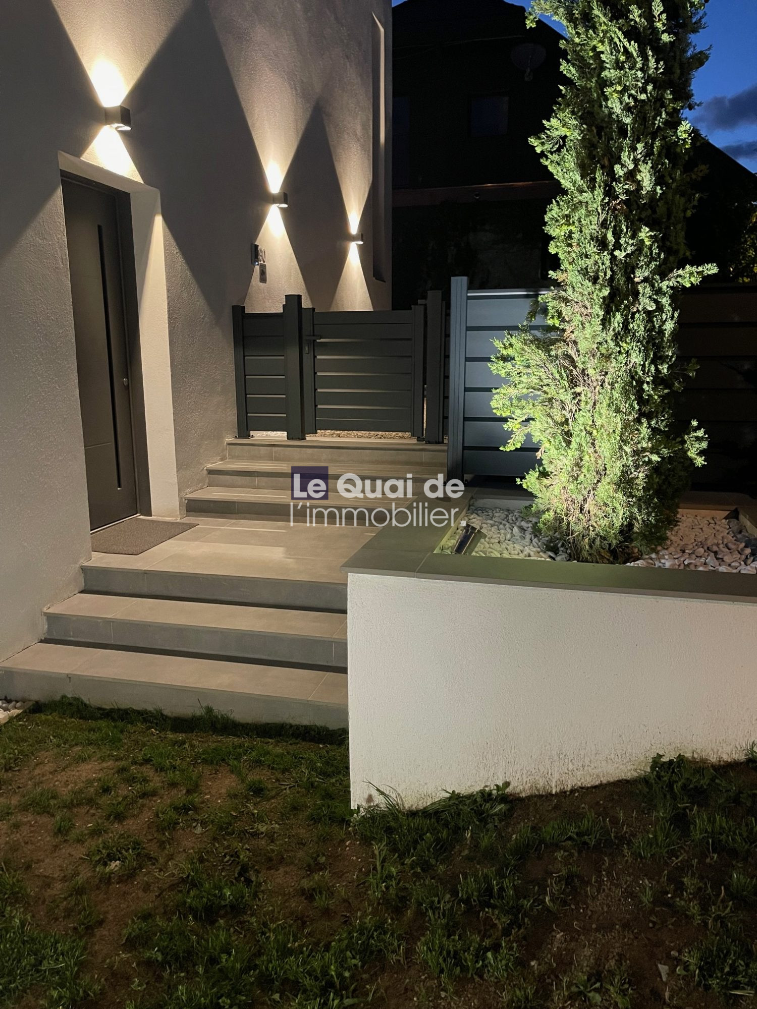 Night exterior entrance: illuminated wall sconces cast triangular light patterns on a gray wall beside a dark door and steps leading to a black gate.