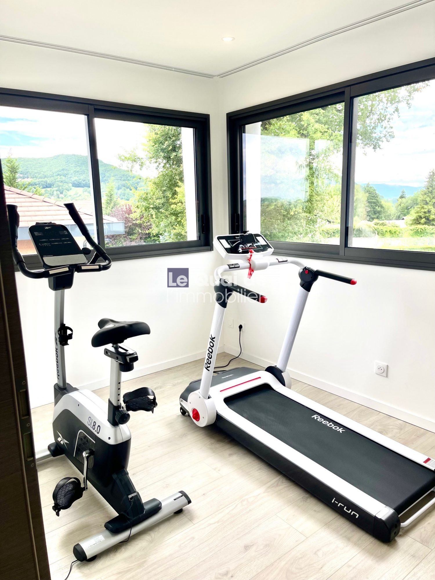 Small home workout room with a white and black treadmill and a white indoor cycling bike by large windows overlooking green hills and trees.