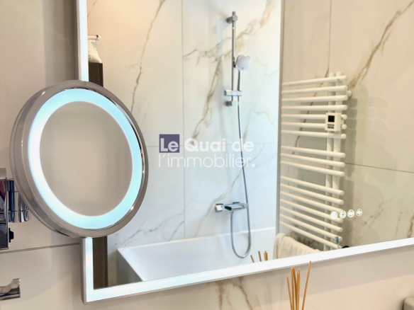 Backlit circular vanity mirror in a marble bathroom with a shower area and white towel warmer visible beside the sink items on the counter.