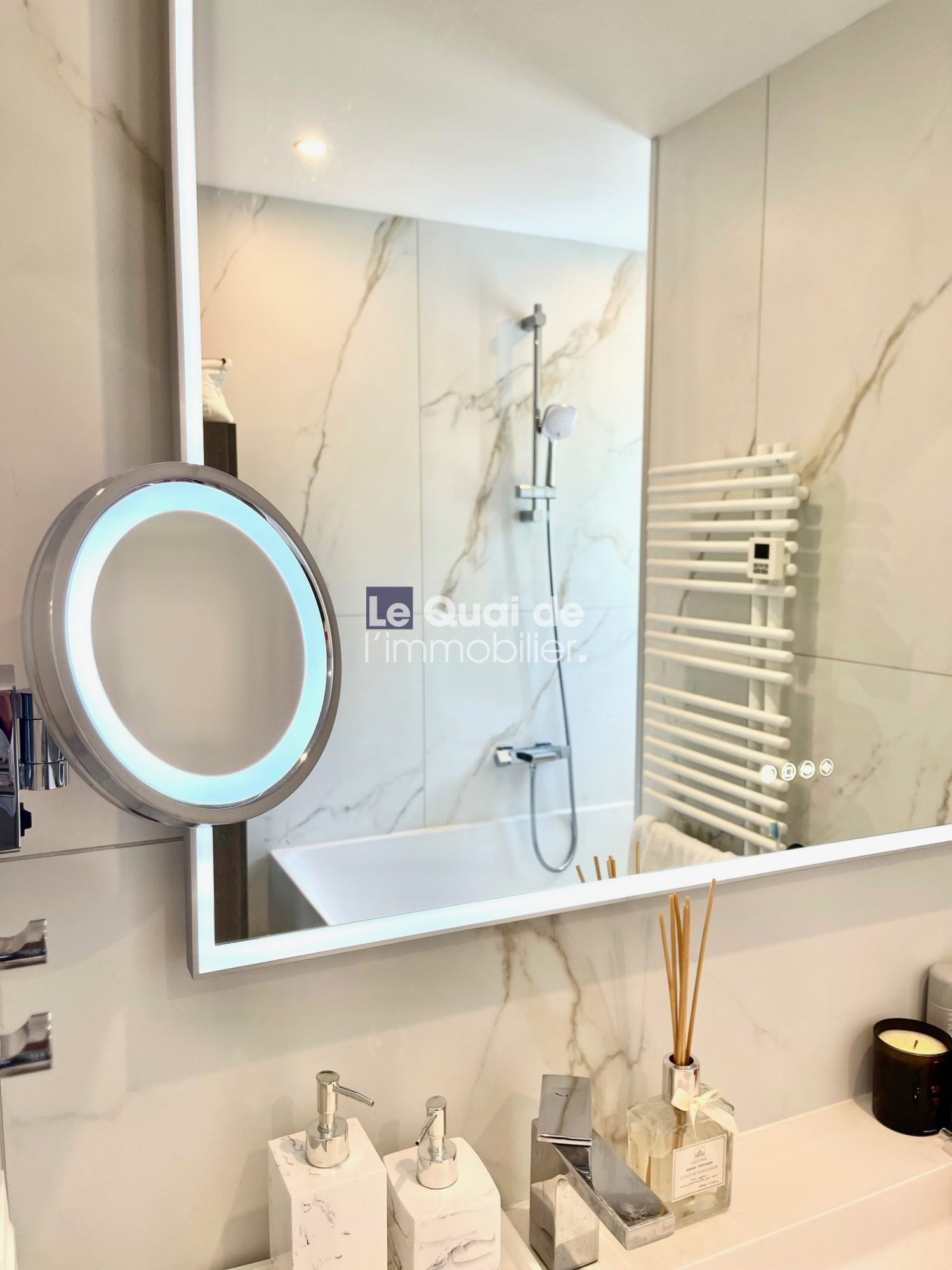 Backlit circular vanity mirror in a marble bathroom with a shower area and white towel warmer visible beside the sink items on the counter.