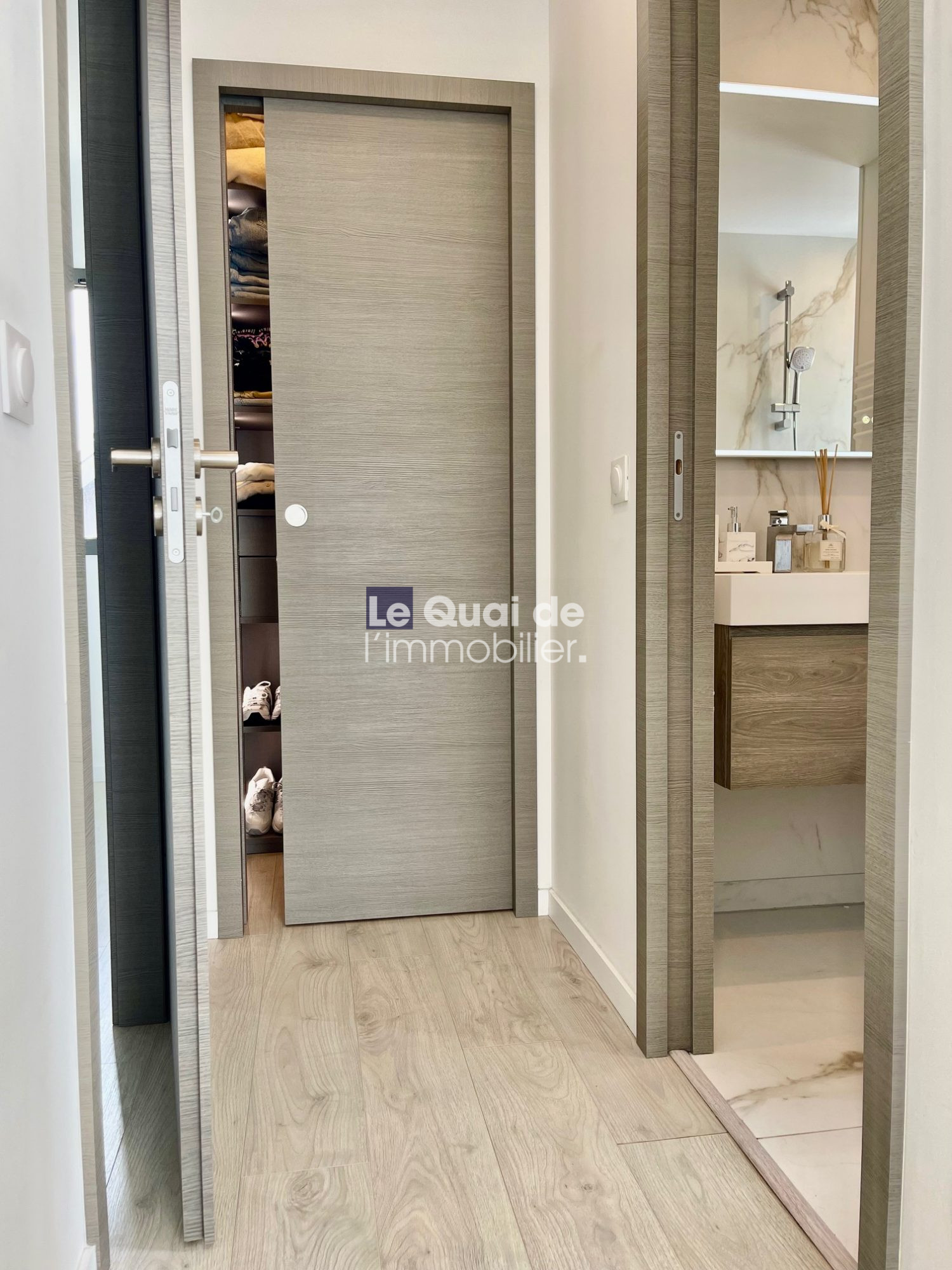 Contemporary hallway view: gray door ajar shows a stacked closet with folded clothes; a nearby doorway reveals a marble-bench bathroom vanity.
