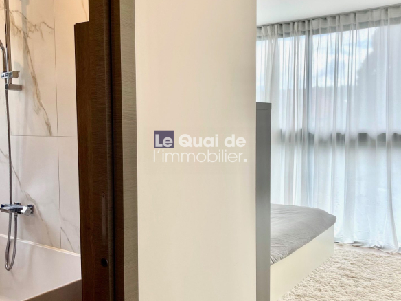 Modern hotel-style suite: bathroom on the left with marble-tiled shower, visible bathtub, and dark wood door frame; bedroom beyond with a low bed and sheer curtains.