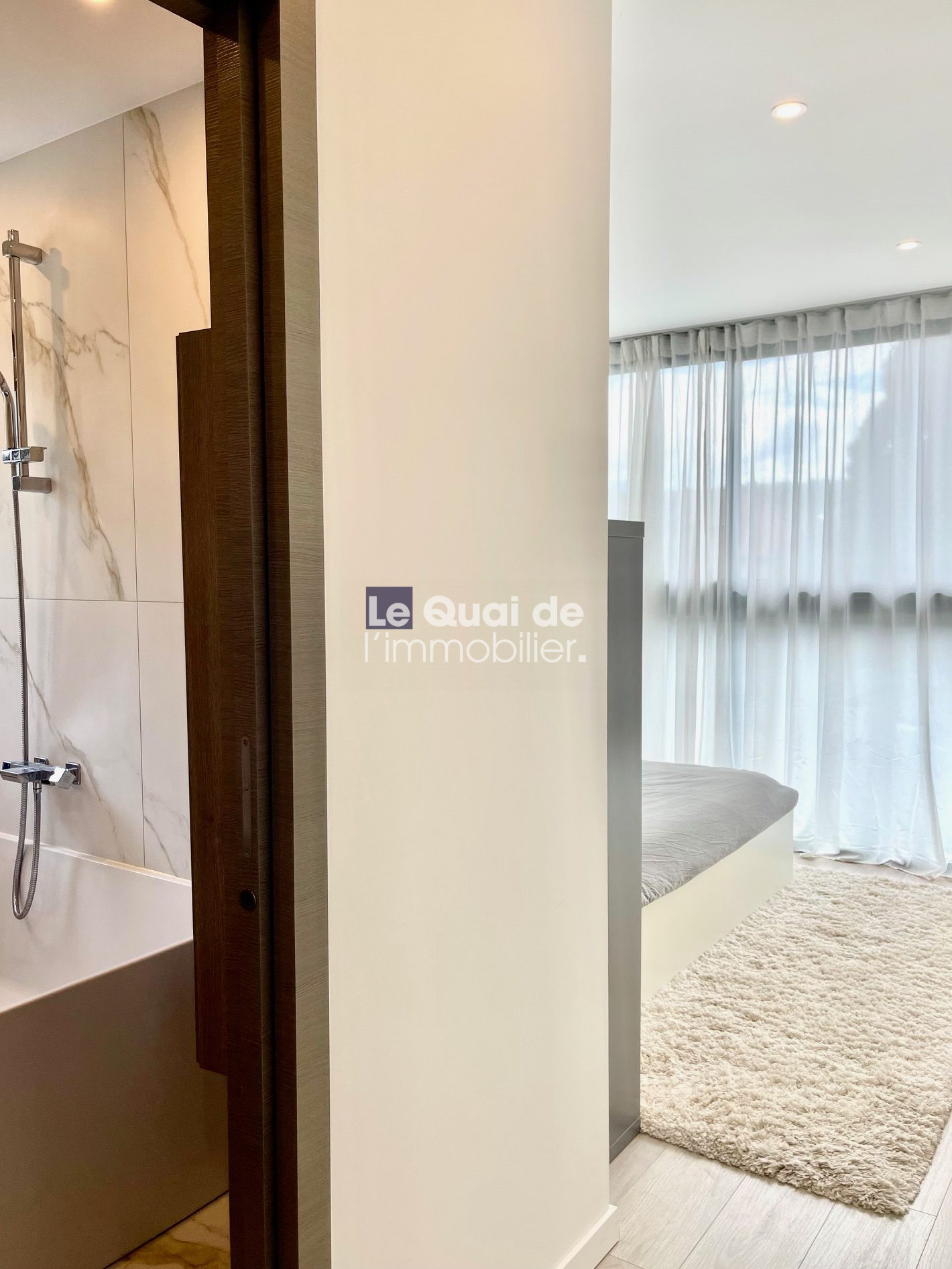 Modern hotel-style suite: bathroom on the left with marble-tiled shower, visible bathtub, and dark wood door frame; bedroom beyond with a low bed and sheer curtains.