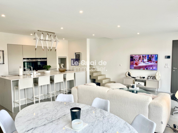 Open-plan kitchen and living area with a white island and six stools, round marble dining table, cream sofa, wall-mounted TV, and concrete stairs.