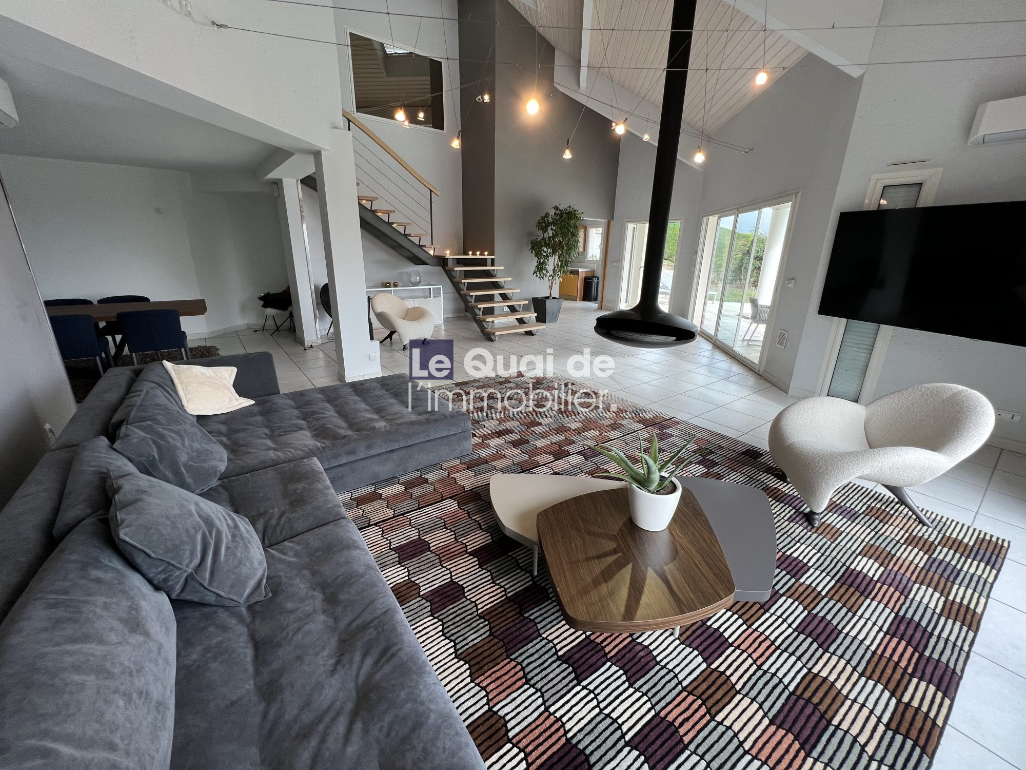 1770966338_VM8231_19_original Open-plan living room with a large gray sectional sofa, a geometric patterned rug, a wooden coffee table with a plant, and a modern white accent chair surrounding a flat-screen TV mounted on the wall.