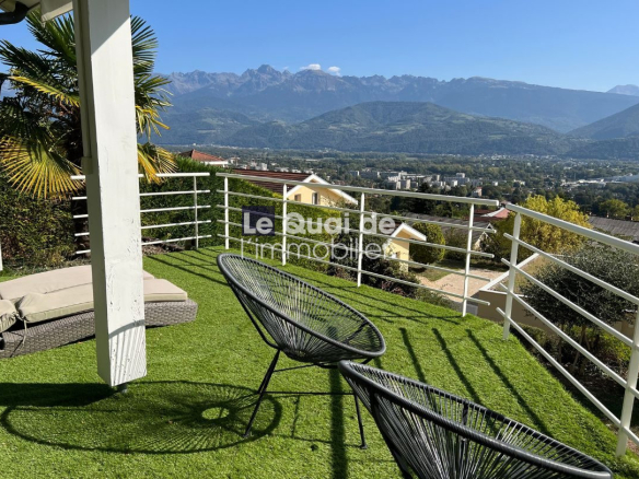 Terrace with artificial grass, two black circular chairs and a beige sofa, white railing, and a mountain valley view on a sunny day.