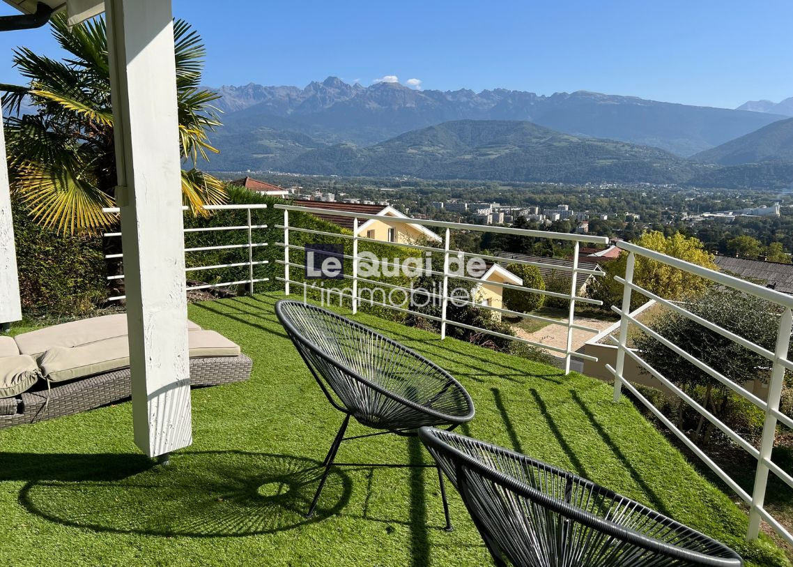 1770966340_VM8231_22_original Terrace with artificial grass, two black circular chairs and a beige sofa, white railing, and a mountain valley view on a sunny day.
