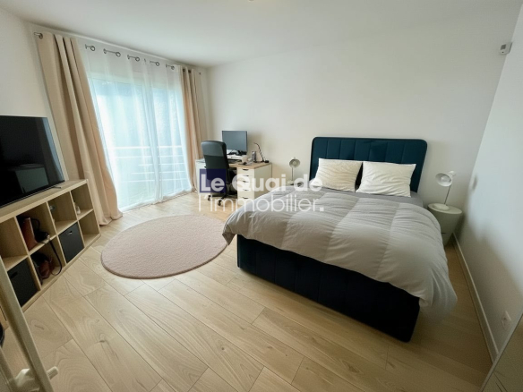 Blue upholstered bed with two white pillows, nightstands with lamps, desk with computer by a large window with beige curtains and light wood floor, circular rug nearby.