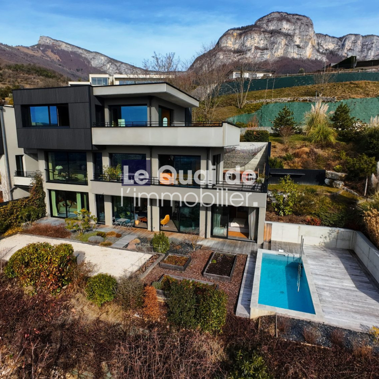 A modern multi-story villa with a rectangular pool, glass doors, and landscaped garden, set against rocky hills in the distance.
