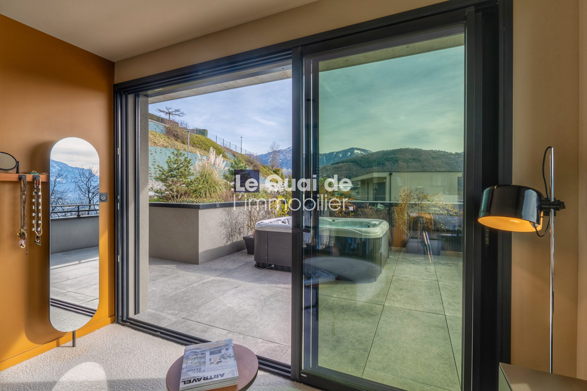 1771603302_VM8233_4_original Modern interior with an orange wall, a tall oval mirror and jewelry hooks on the left, sliding glass doors opening to a terrace with a hot tub and mountain views outside.