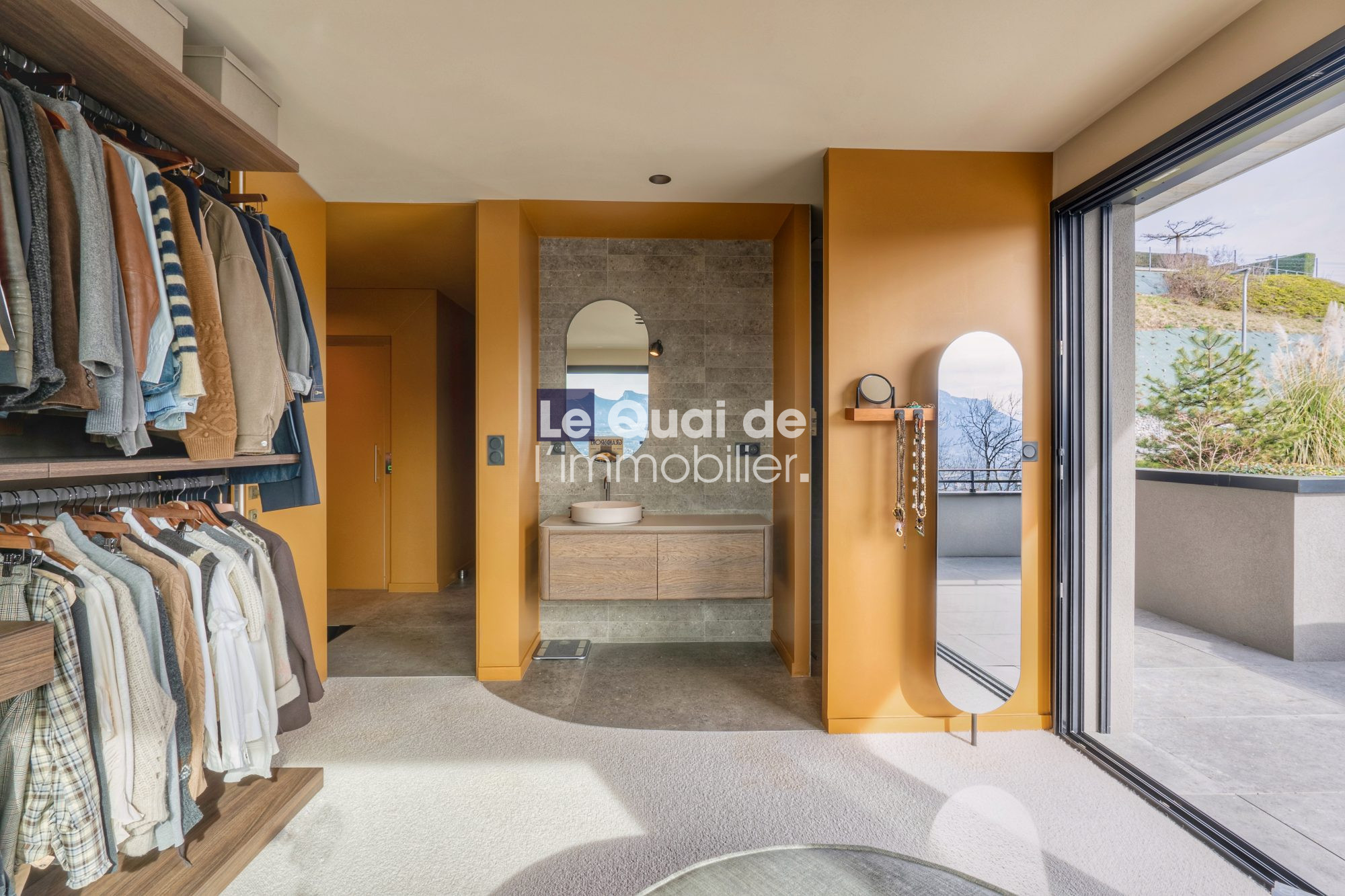 1771603303_VM8233_5_original Walk-in closet with orange walls, clothes on racks to the left, a wooden vanity with a round sink in the center, and a glass door opening to an outdoor terrace on the right.