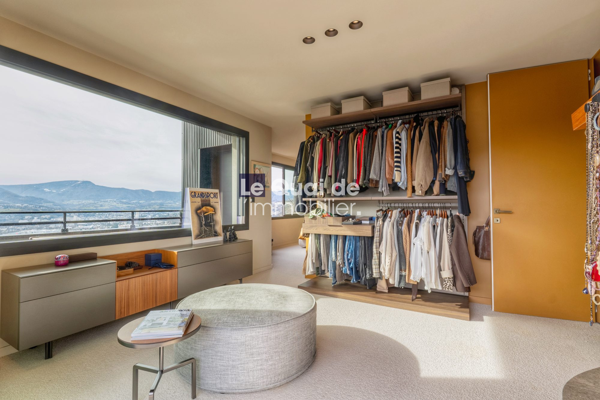 1771603307_VM8233_7_original Spacious walk-in closet with double hanging racks full of clothes, a low cabinet, ottoman, and a window with mountain view.