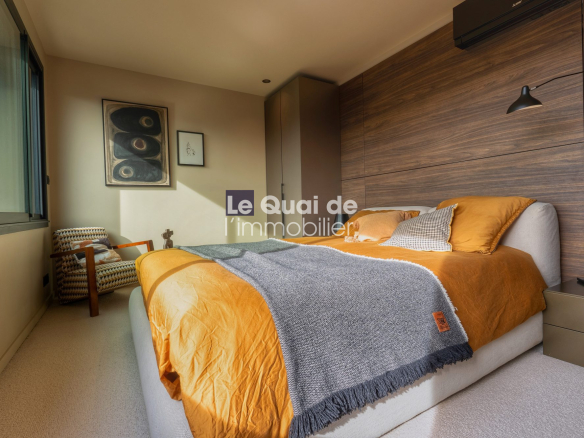 Modern bedroom with an orange duvet, gray throw, and wooden paneled headboard wall; a beige upholstered bed and side table sit beside a large window and a patterned chair in the corner.