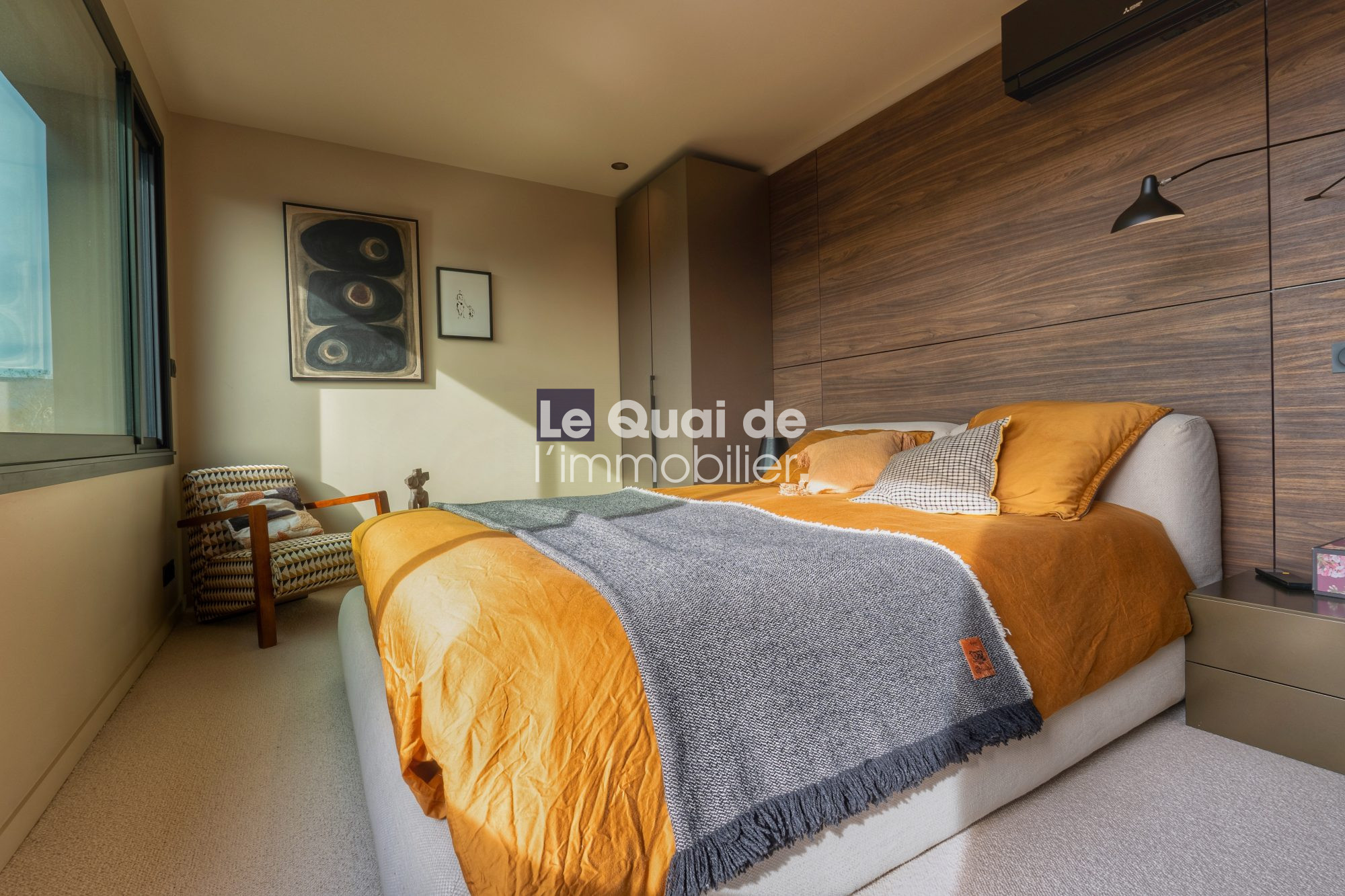 1771603308_VM8233_8_original Modern bedroom with an orange duvet, gray throw, and wooden paneled headboard wall; a beige upholstered bed and side table sit beside a large window and a patterned chair in the corner.