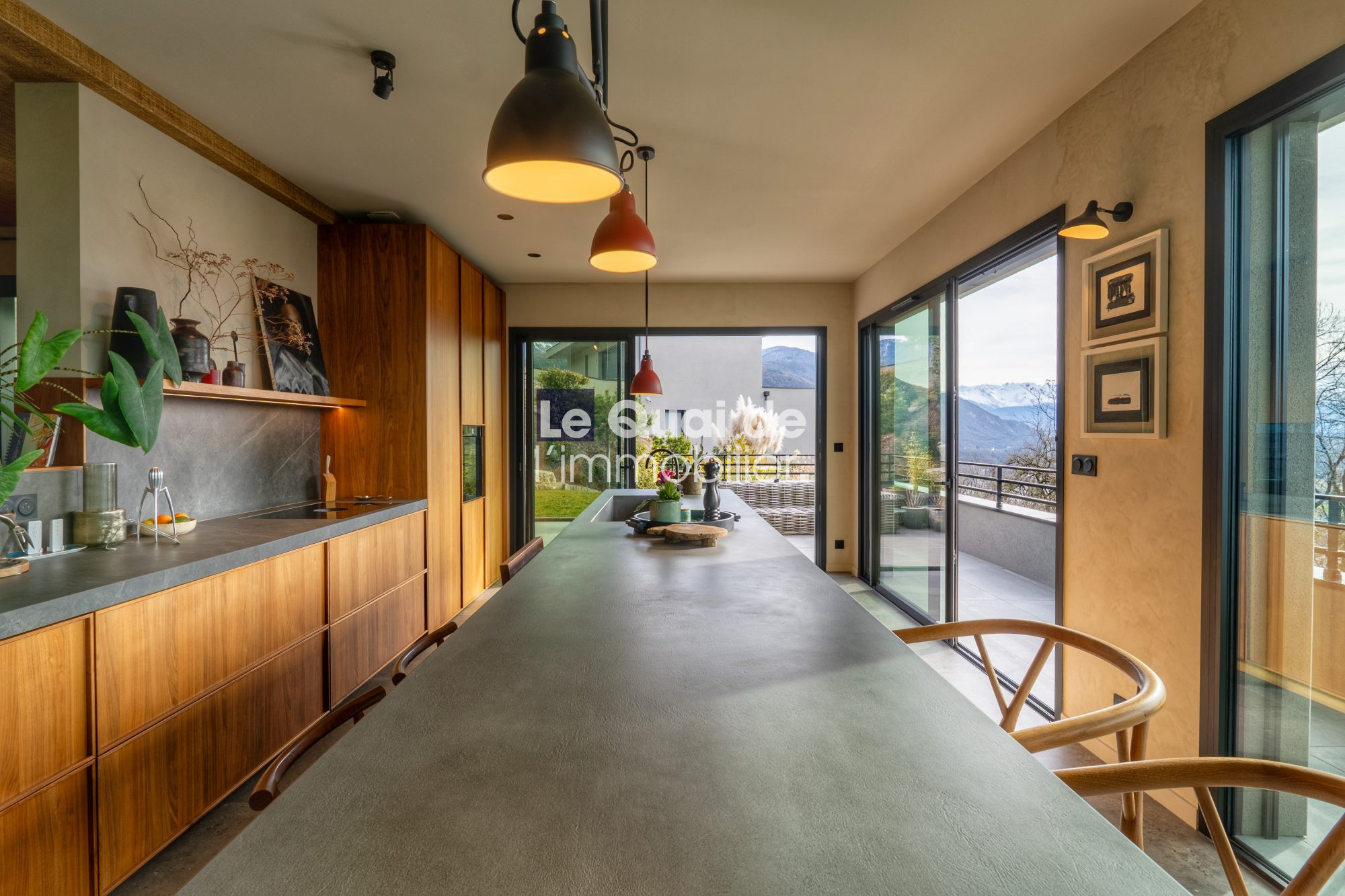 1771603313_VM8233_11_original Modern kitchen with a long concrete island, warm wooden cabinets, and sliding glass doors to a mountain view outside.
