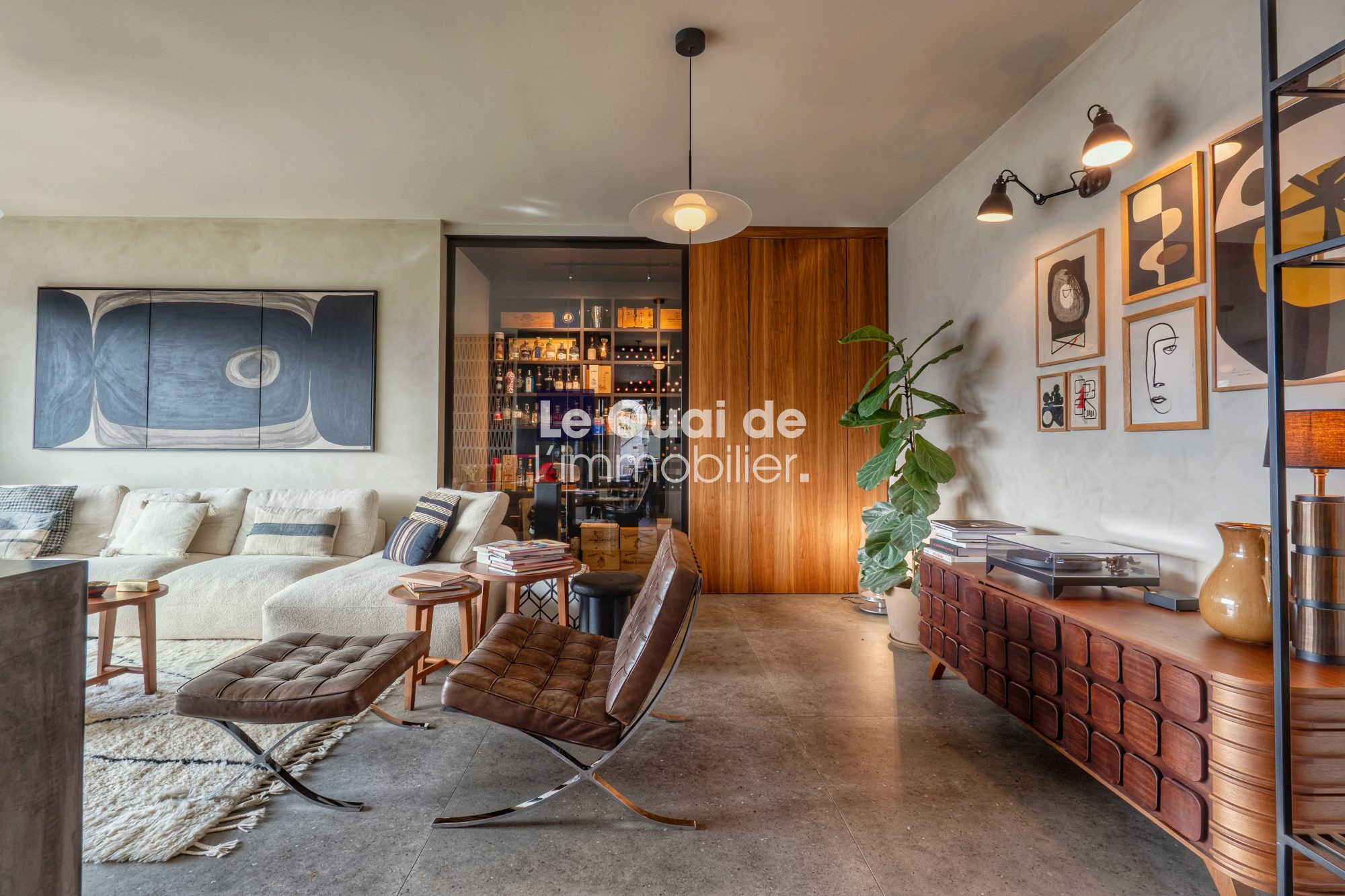 1771603316_VM8233_13_original Modern living room with a beige sectional, brown leather lounge chair and ottoman, and a wooden sideboard; gallery wall and a tall plant contribute to a cozy, stylish space.