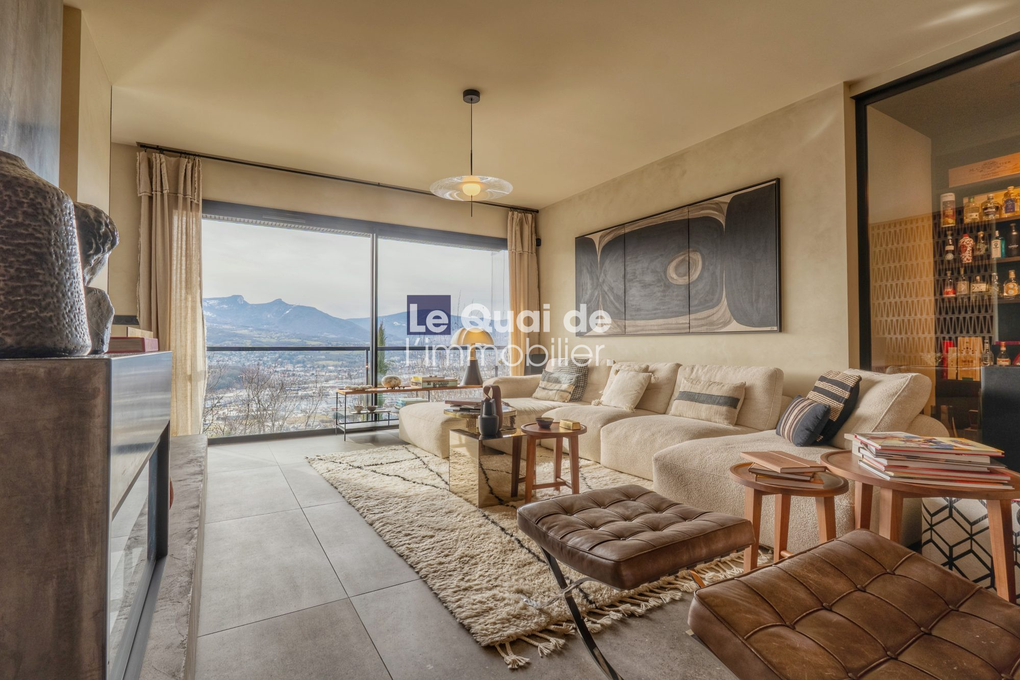 1771603317_VM8233_14_original Bright living room with a beige sectional, cream rug, and a large window showing mountains and a lake outside.