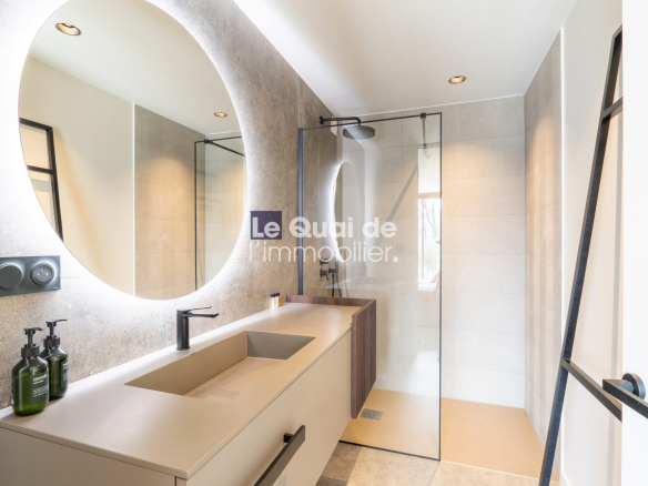 Circular backlit mirror over a beige double vanity with integrated sink, in a modern bathroom with a glass shower enclosure and black fixtures.