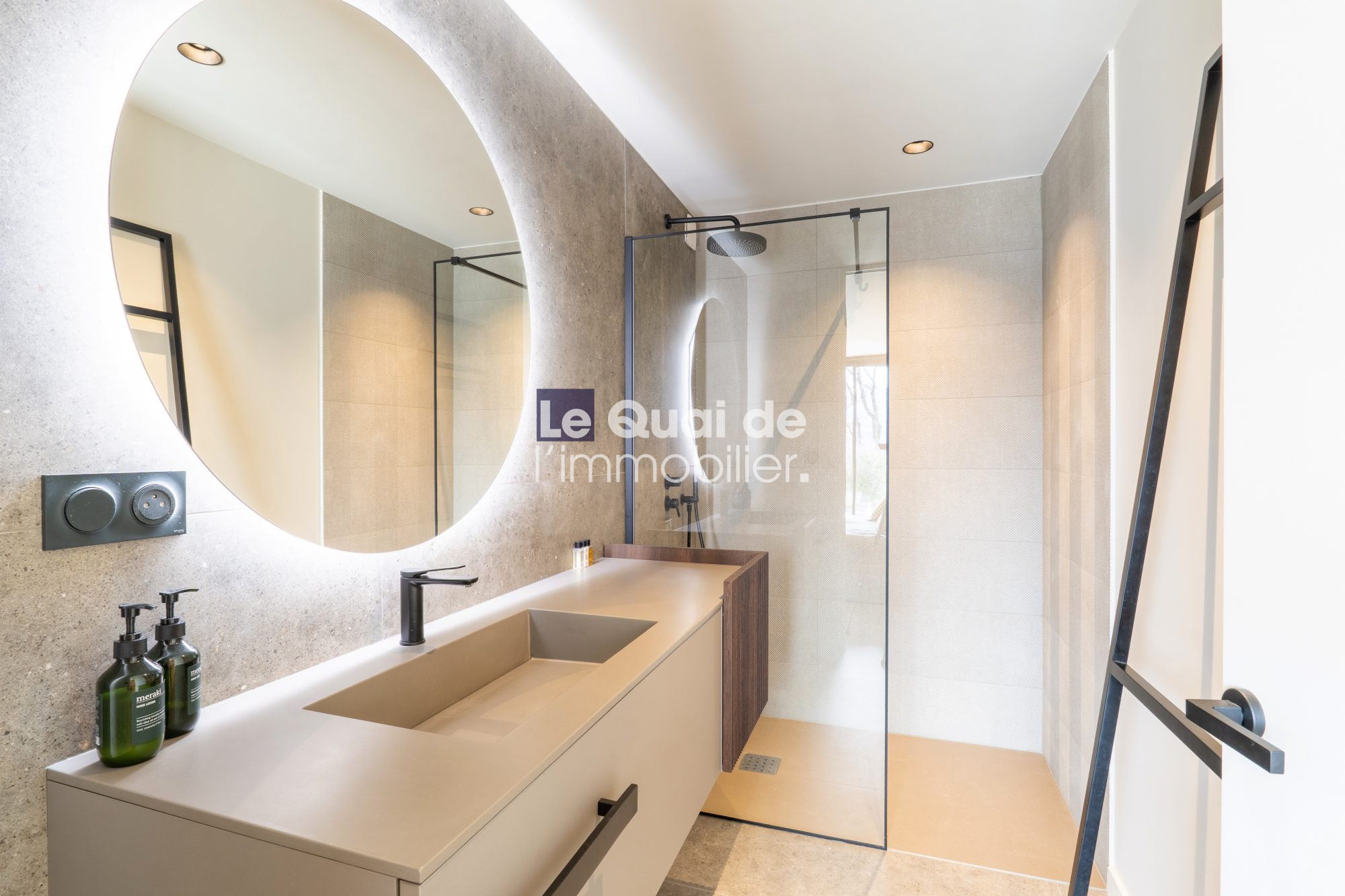 1771603335_VM8233_27_original Circular backlit mirror over a beige double vanity with integrated sink, in a modern bathroom with a glass shower enclosure and black fixtures.