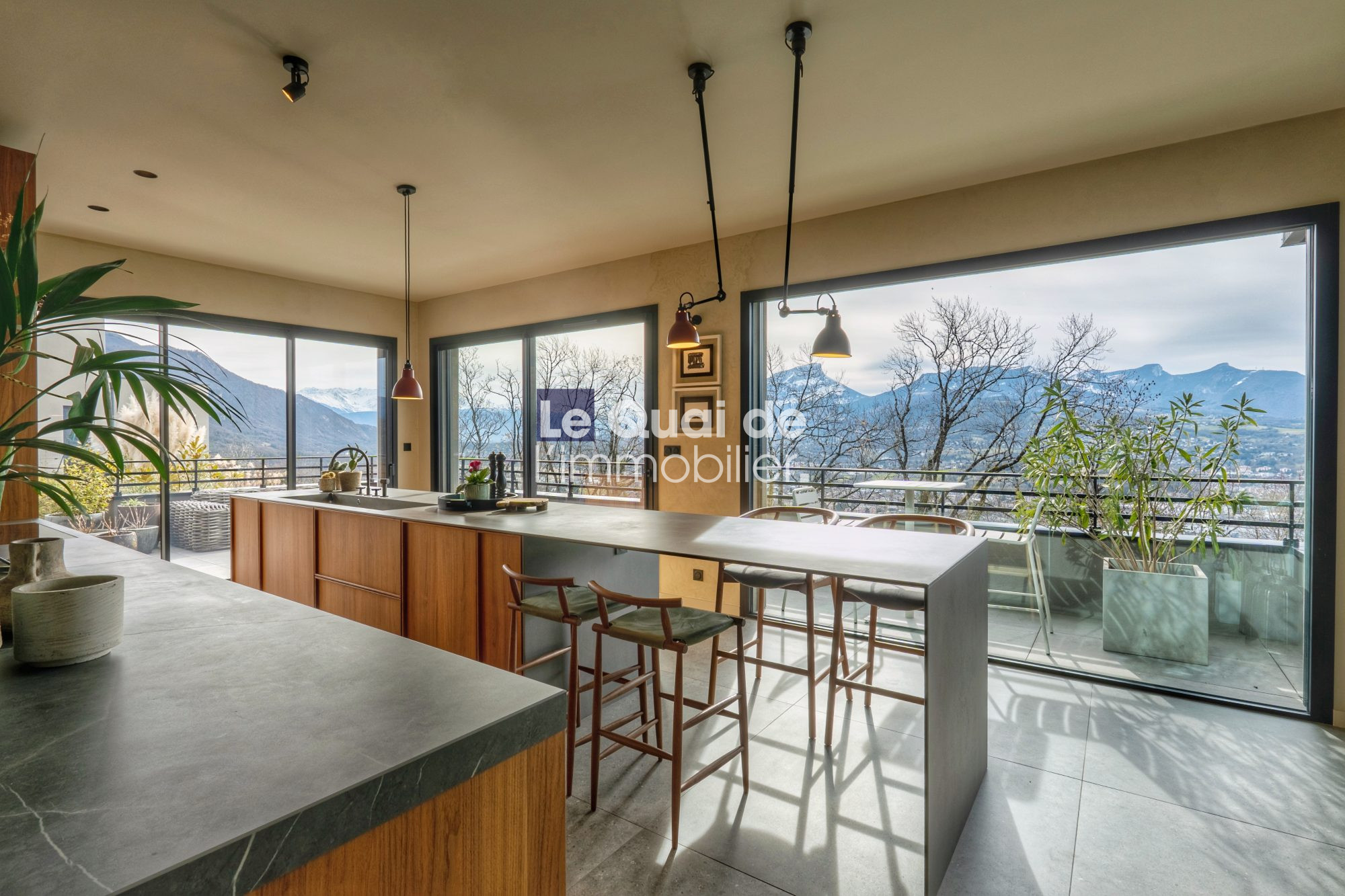 1771603338_VM8233_29_original Modern kitchen with a long island, wooden cabinets, and large windows showing a mountain landscape outside (open-plan dining area).