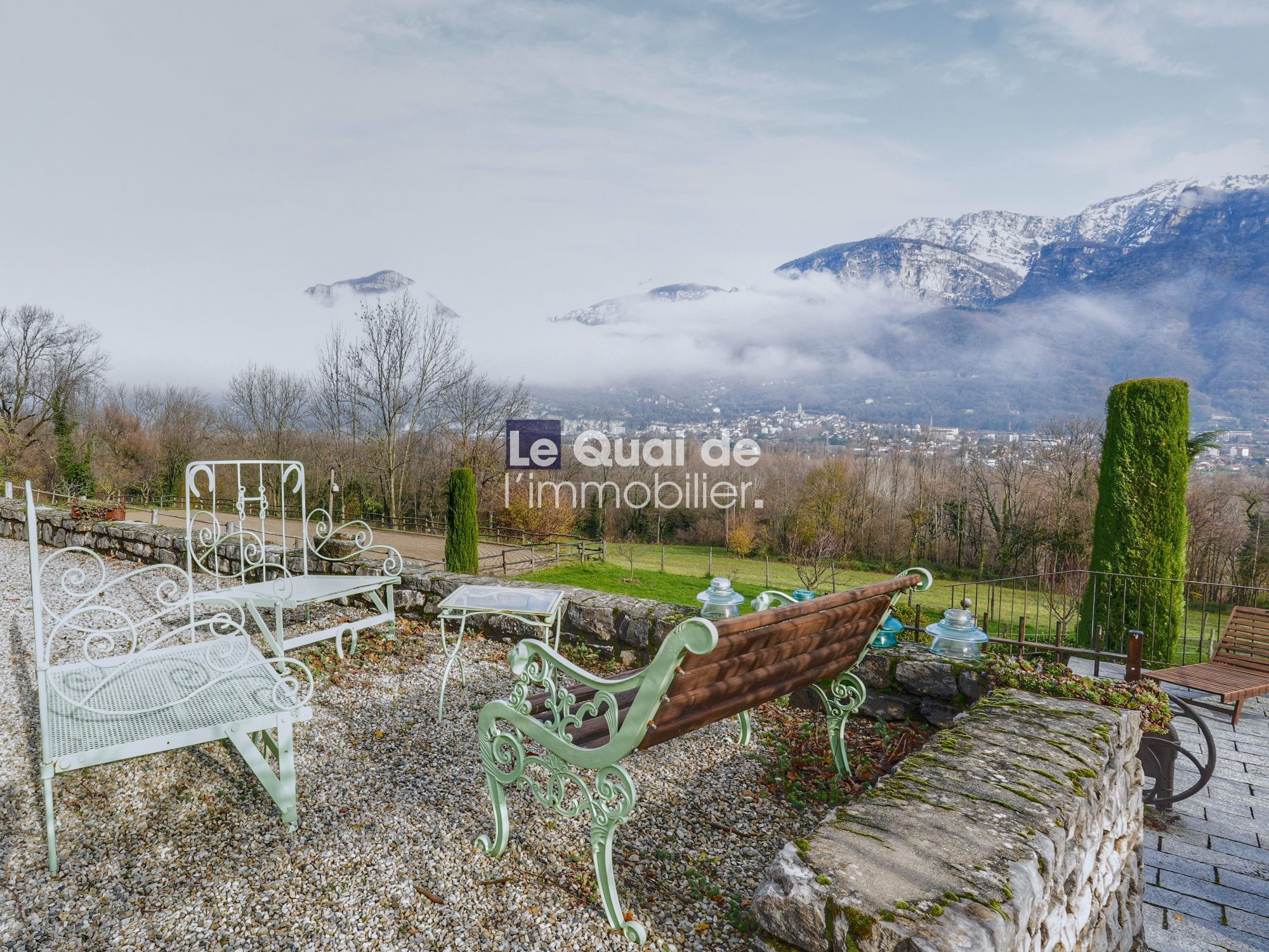 Garden seating area with ornate white and green benches facing a valley and snow-capped mountains in the distance under a cloudy sky.