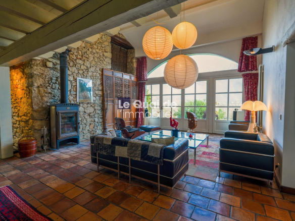 Cozy living room with a stone wall and wood-burning stove, blue leather sofas, glass coffee table, and large arched windows with red curtains.