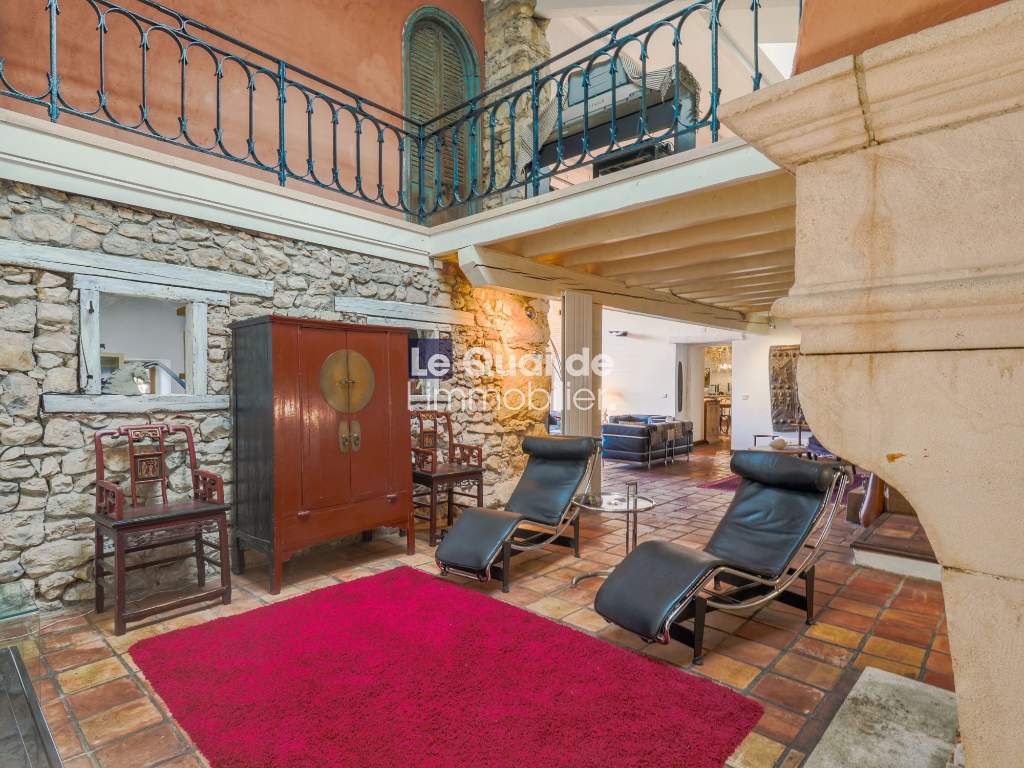 Rustic indoor-outdoor living area with stone walls, blue railing above, a red wardrobe, wooden chairs, black leather lounge chairs, and a bright red rug on terracotta tiles.