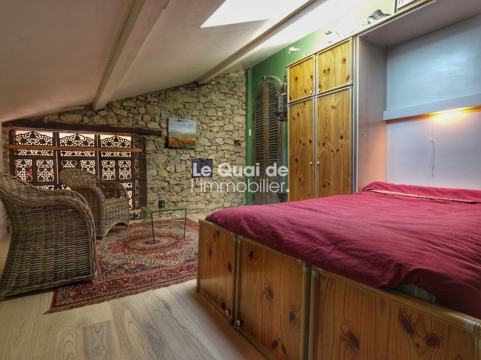 Loft bedroom with a stone wall, wicker chairs, glass coffee table, and a red bedspread next to a tall wooden wardrobe.