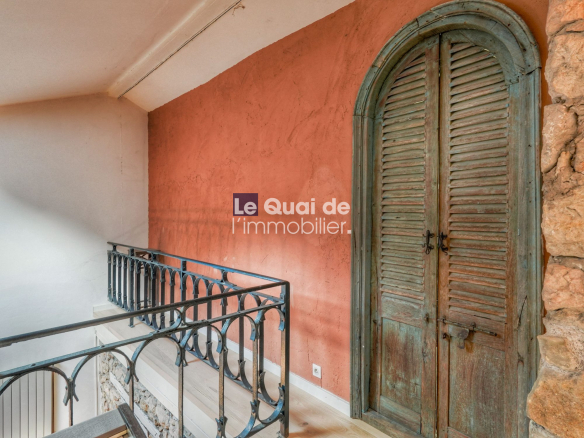 Terracotta-colored wall with a tall arched, weathered teal wooden door beside a stone wall and decorative iron railing; watermark 'Le Quai de l’immobilier'.