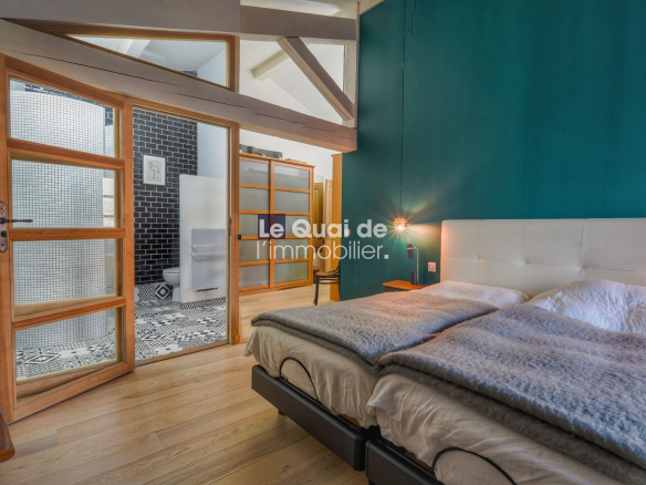 Bedroom with teal accent wall, white upholstered bed and gray throws; glass-panel wooden doors open to a bathroom with patterned black-and-white tiles.