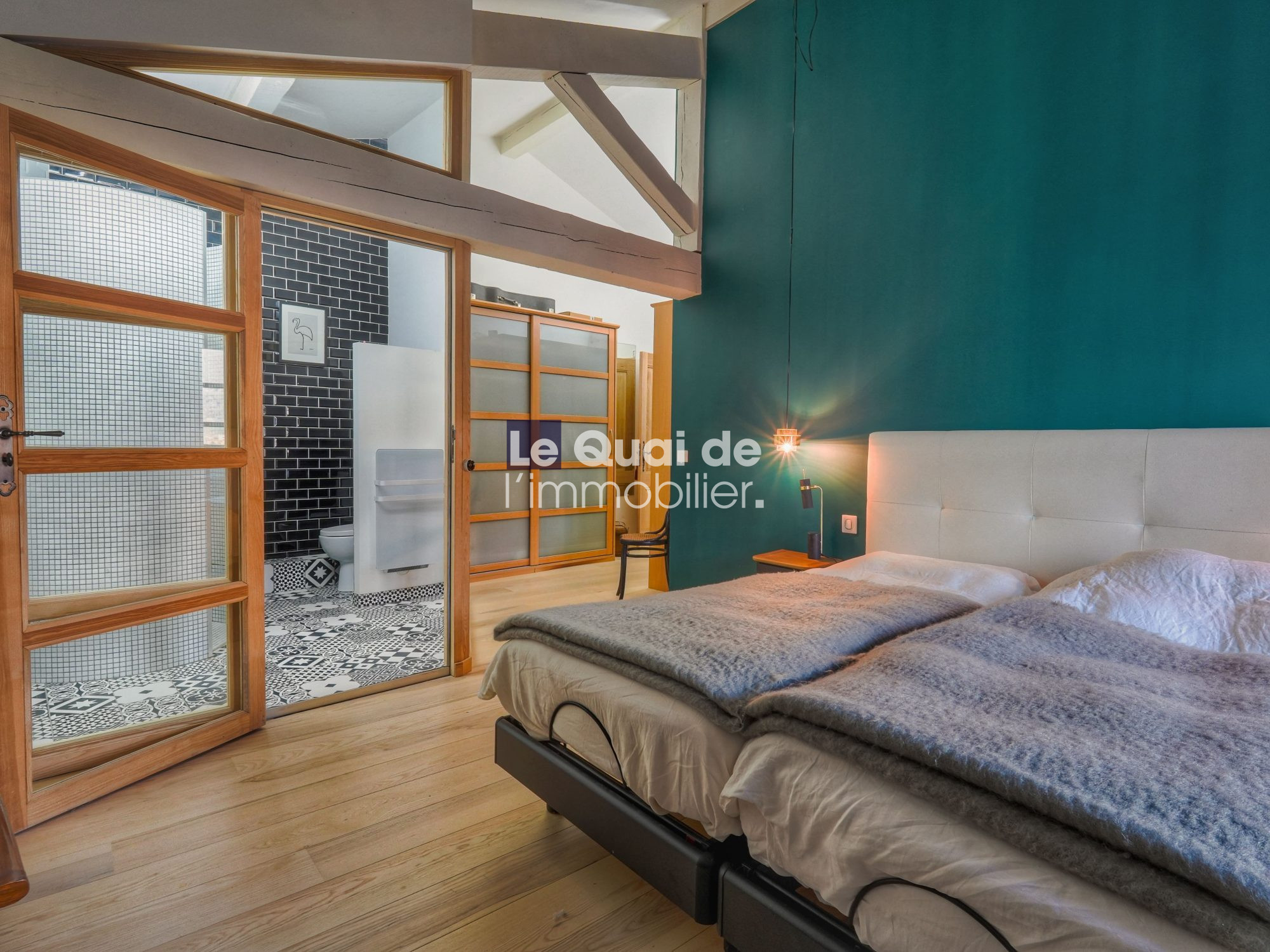 Bedroom with teal accent wall, white upholstered bed and gray throws; glass-panel wooden doors open to a bathroom with patterned black-and-white tiles.