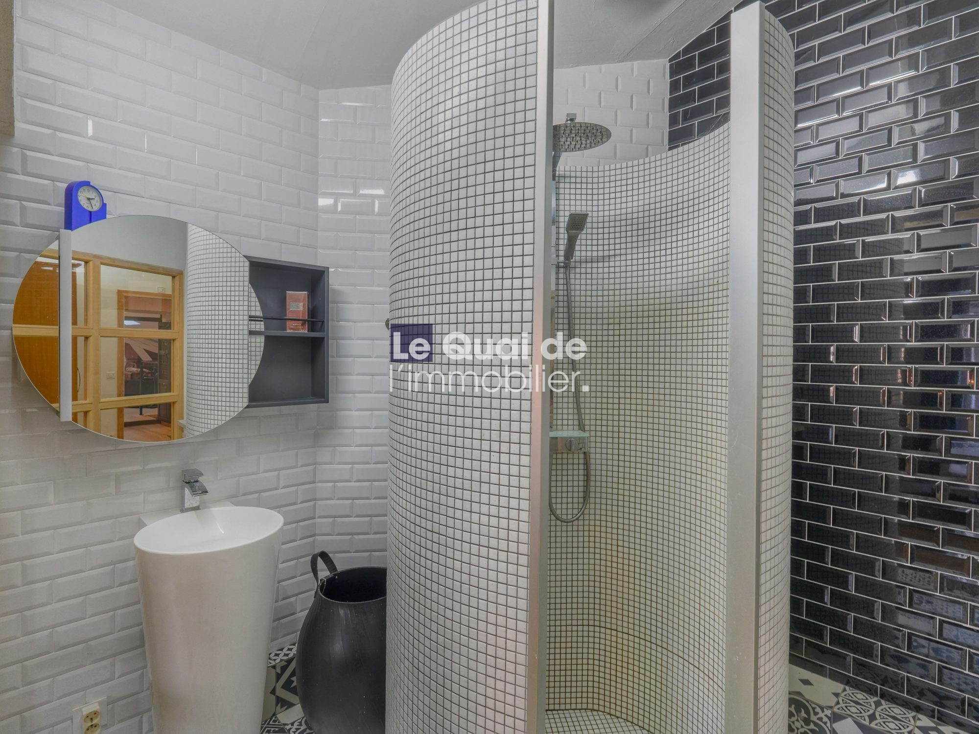 Showroom bathroom with a curved mosaic-tiled shower enclosure, round mirror, and white pedestal sink on a patterned floor.
