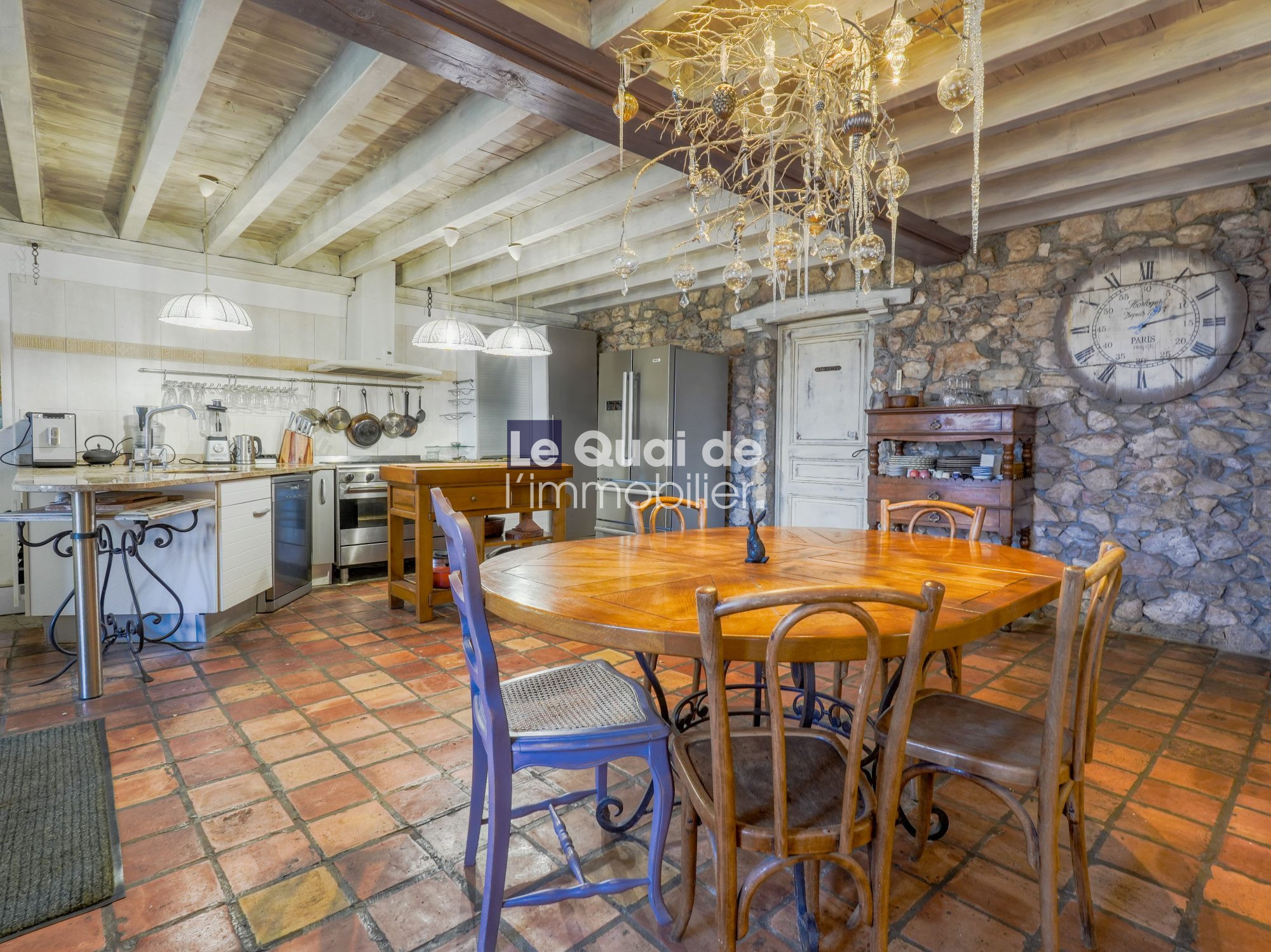 Rustic kitchen and dining area with a round wooden table, mismatched chairs, stone wall, and a large clock.