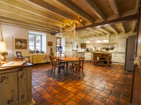 Rustic kitchen and dining area with a round wooden table, chairs, exposed wooden beams, and white cabinets in the background under warm lighting.