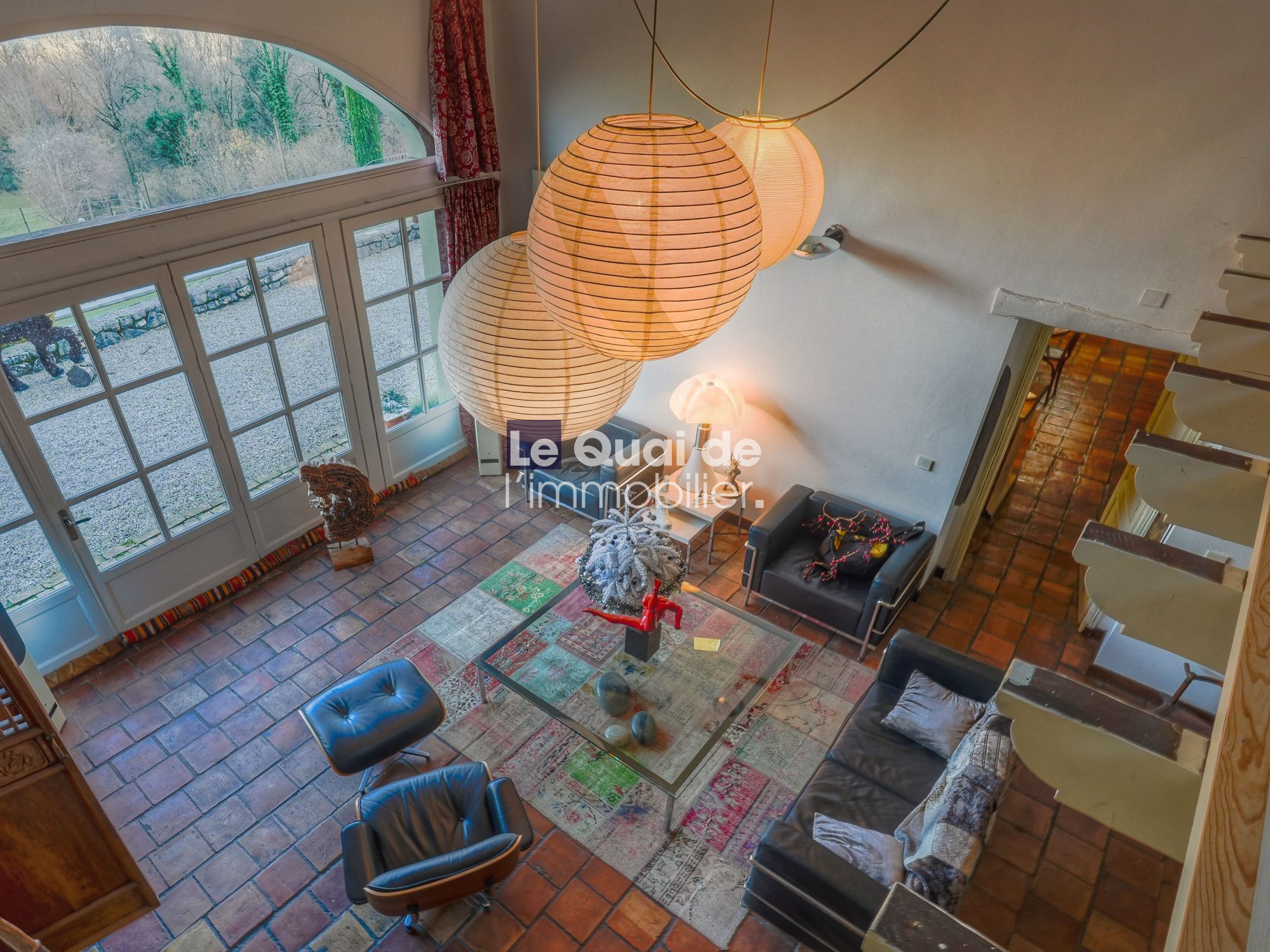 Bright living room viewed from above: terracotta tile floor, glass coffee table, black leather sofas, colorful rug, large arched windows/doors to a garden, and three round paper lantern pendant lights; curved staircase on the right.
