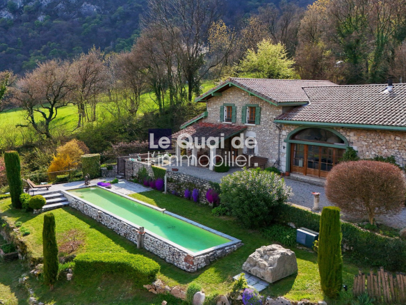 Stone villa with tiled roof and green-shuttered windows, surrounded by a manicured garden and a long rectangular pool. A rural hillside backdrop.