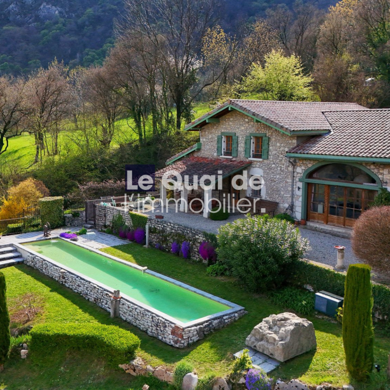 Stone villa with tiled roof and green-shuttered windows, surrounded by a manicured garden and a long rectangular pool. A rural hillside backdrop.