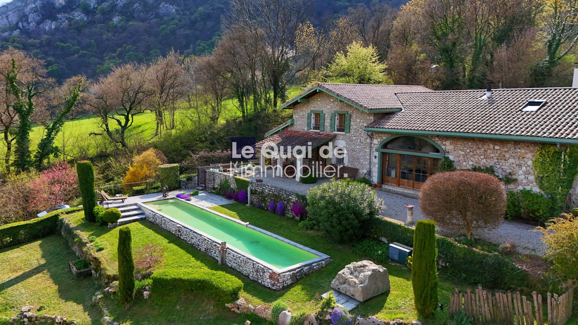 Stone villa with tiled roof and green-shuttered windows, surrounded by a manicured garden and a long rectangular pool. A rural hillside backdrop.