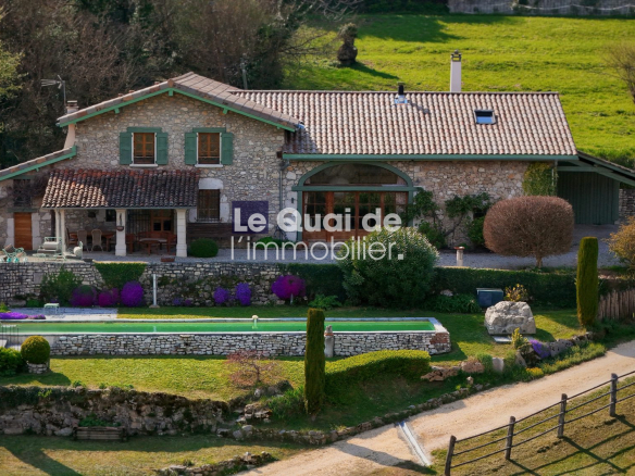 Stone cottage with tiled roof and green shutters, surrounded by a landscaped garden and a rectangular pool in the countryside.