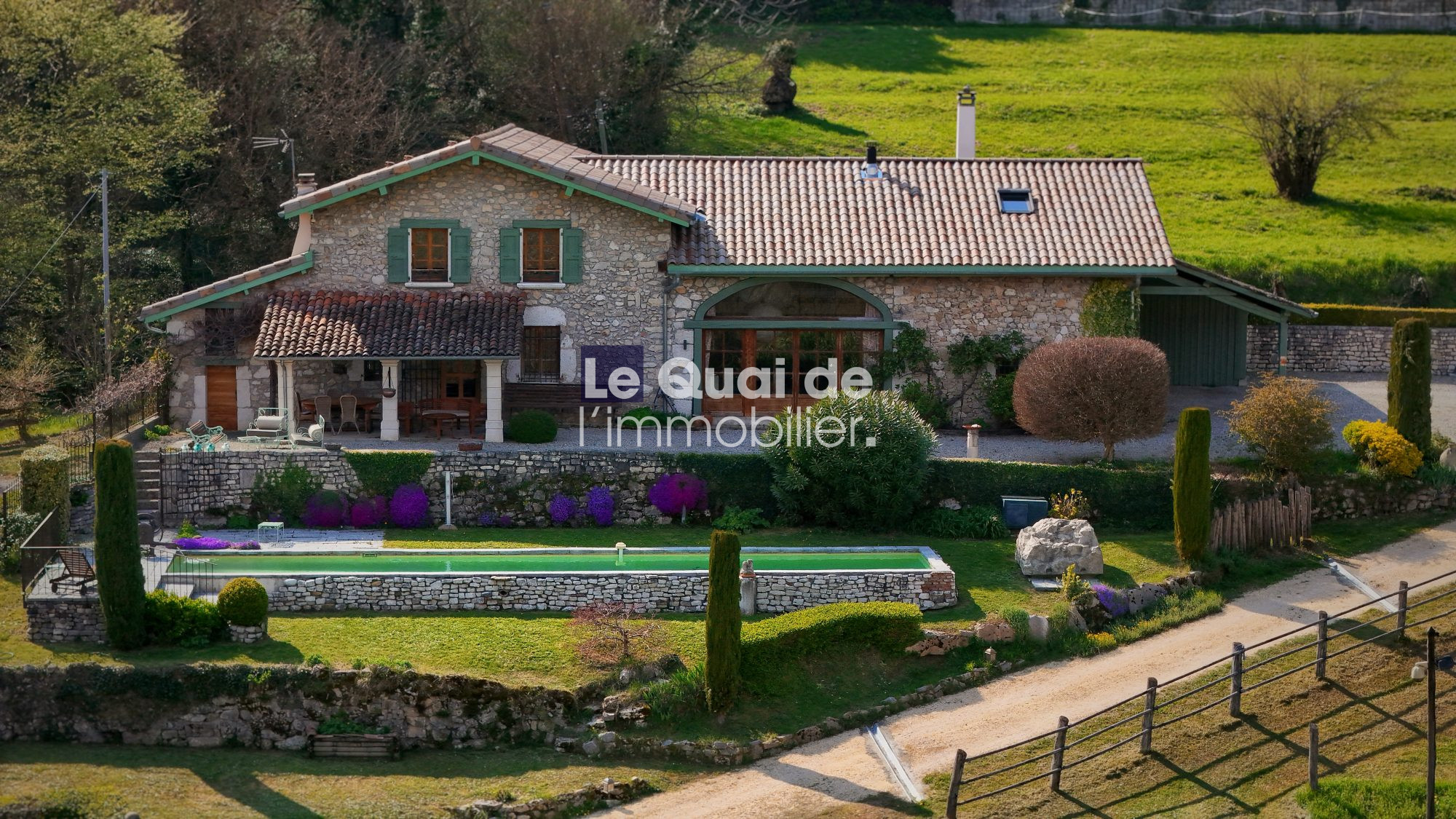 Stone cottage with tiled roof and green shutters, surrounded by a landscaped garden and a rectangular pool in the countryside.