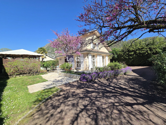 Two-story beige house with white shutters, surrounded by blooming pink tree and purple flowers on a sunny day