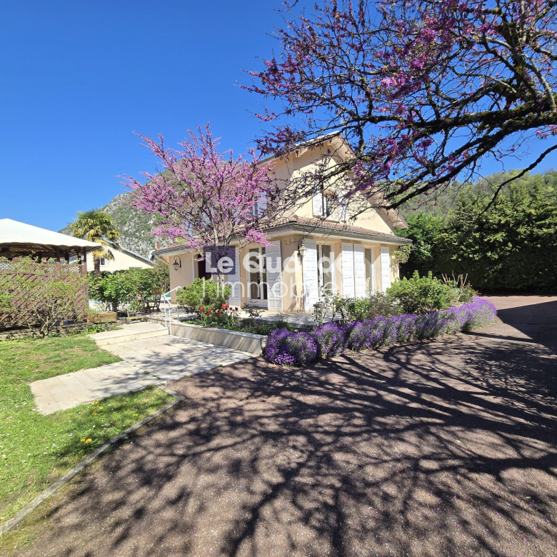 Two-story beige house with white shutters, surrounded by blooming pink tree and purple flowers on a sunny day