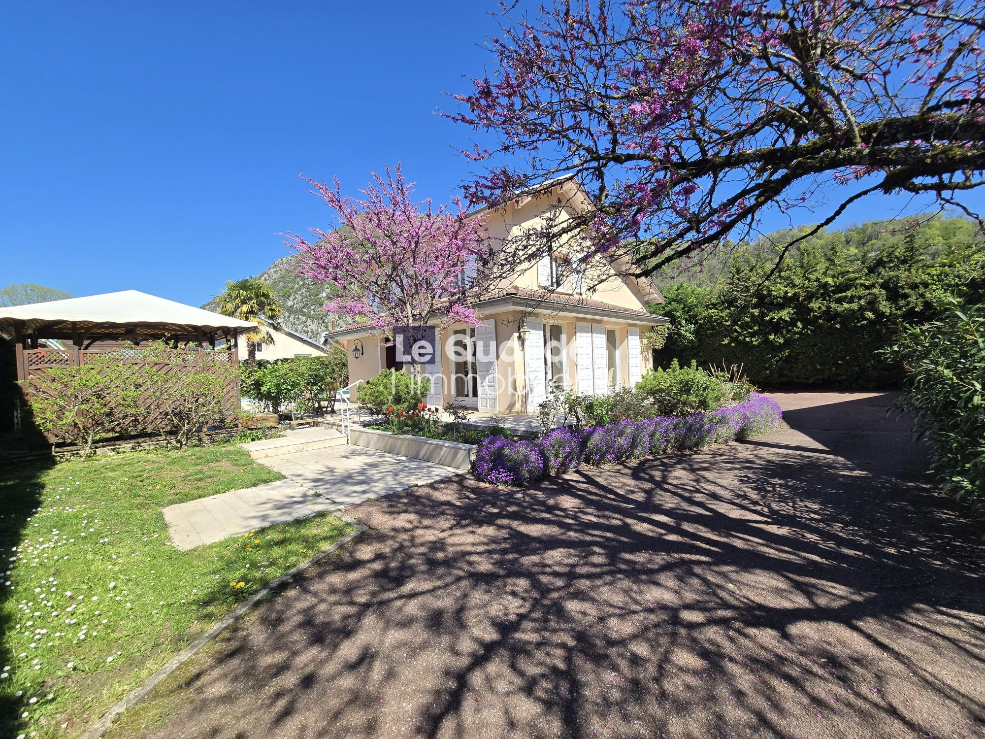 Two-story beige house with white shutters, surrounded by blooming pink tree and purple flowers on a sunny day