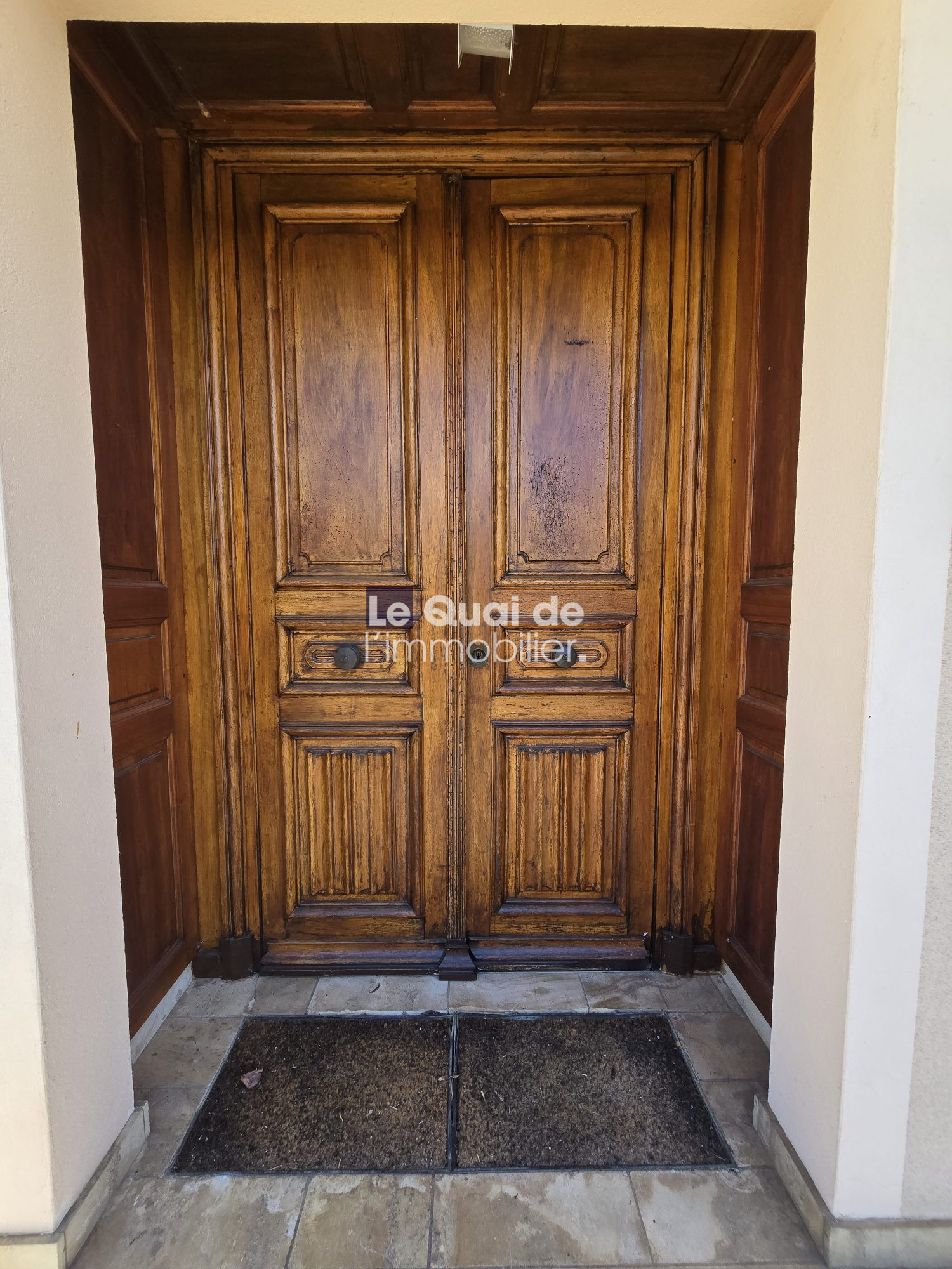 Double tall wooden doors with paneled carvings at a building entrance; a tiled threshold and a dirt mat in front.