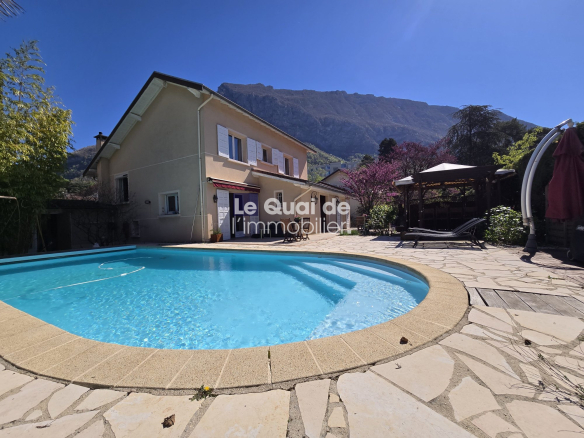 Bright outdoor scene: a kidney-shaped pool in a stone patio with a beige house and mountain backdrop; watermark reads 'Le Quai de l'immobilier'.