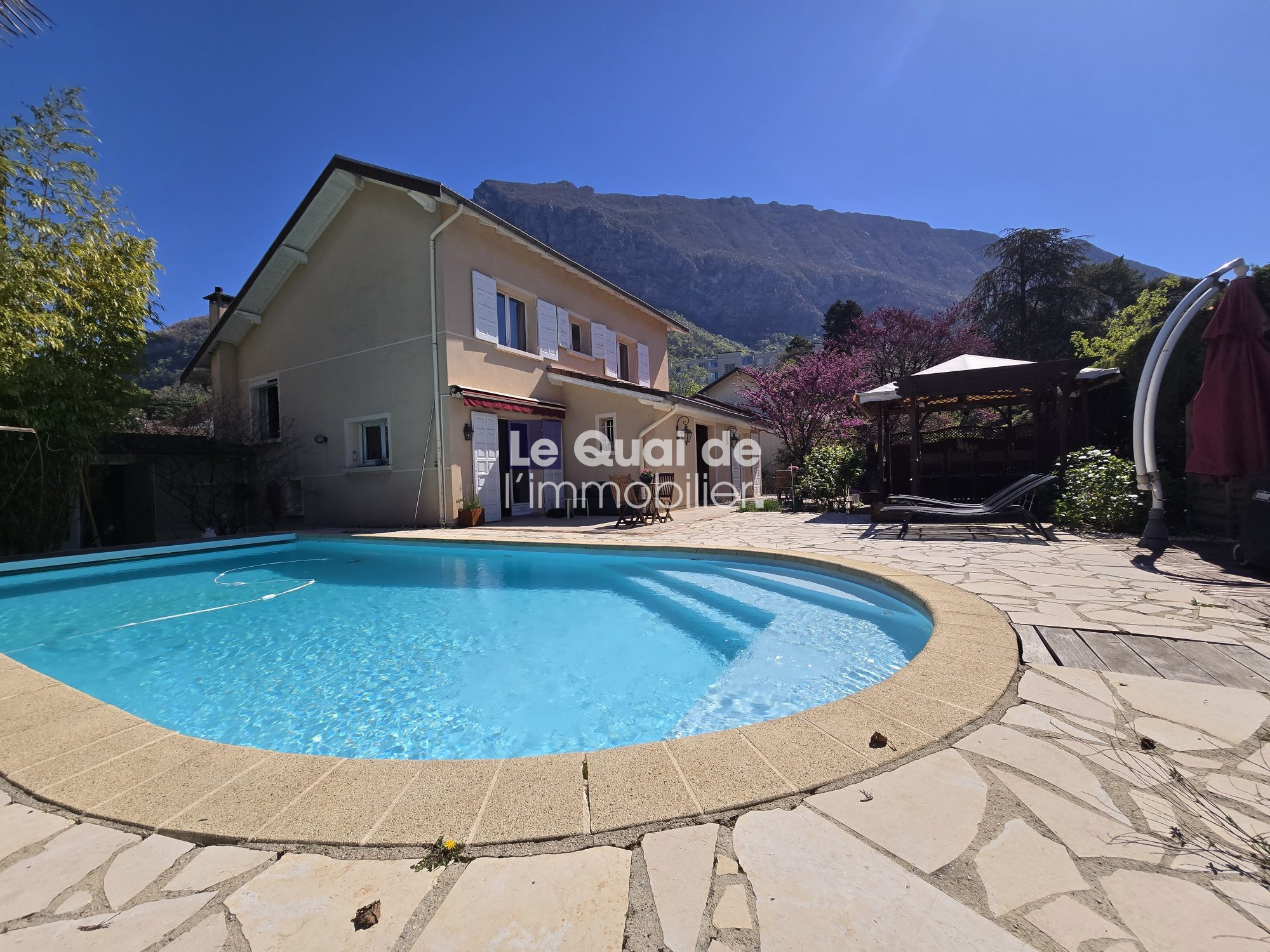 Bright outdoor scene: a kidney-shaped pool in a stone patio with a beige house and mountain backdrop; watermark reads 'Le Quai de l'immobilier'.