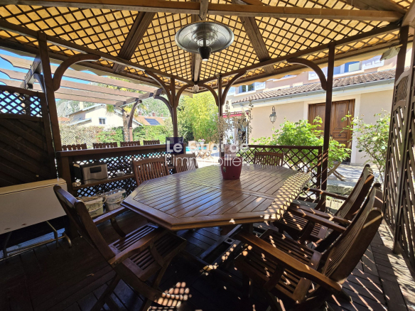 Outdoor wooden patio with a large dining table and many chairs under a lattice pergola, sunlight and greenery in the yard.