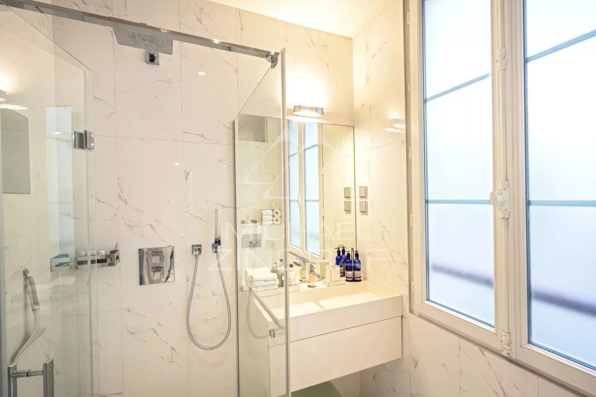 Salle de bain moderne avec douche en verre et lavabo, lumière naturelle à travers une fenêtre.